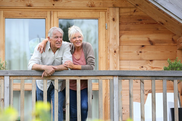 An older couple stands on the wooden balcony of a cabin, smiling and looking into the distance. The woman has her arm around the mans shoulders. The cabin is made of light-colored wood with large windows.