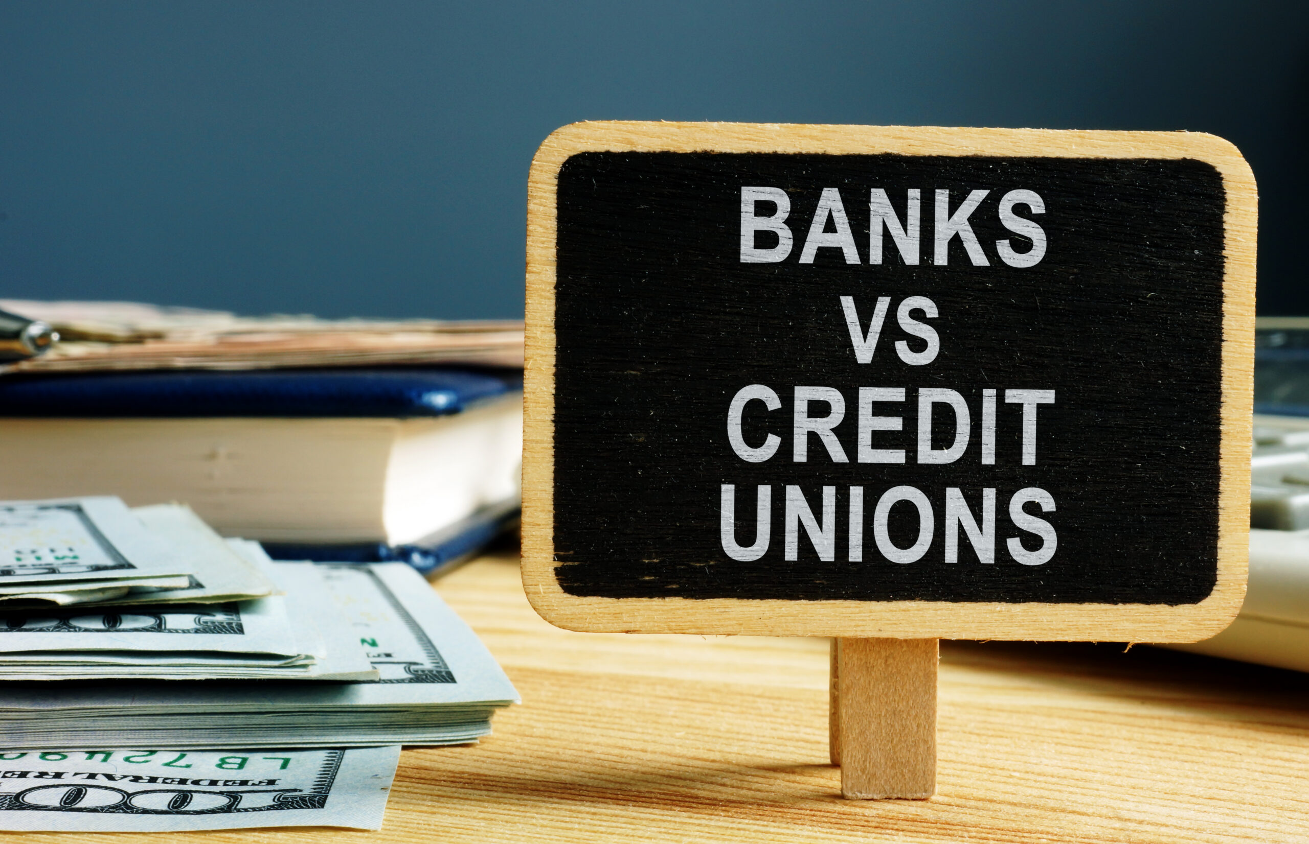 A small chalkboard sign reading Banks vs Credit Unions stands on a desk next to stacks of cash, a notebook, and a calculator, symbolizing a financial comparison.