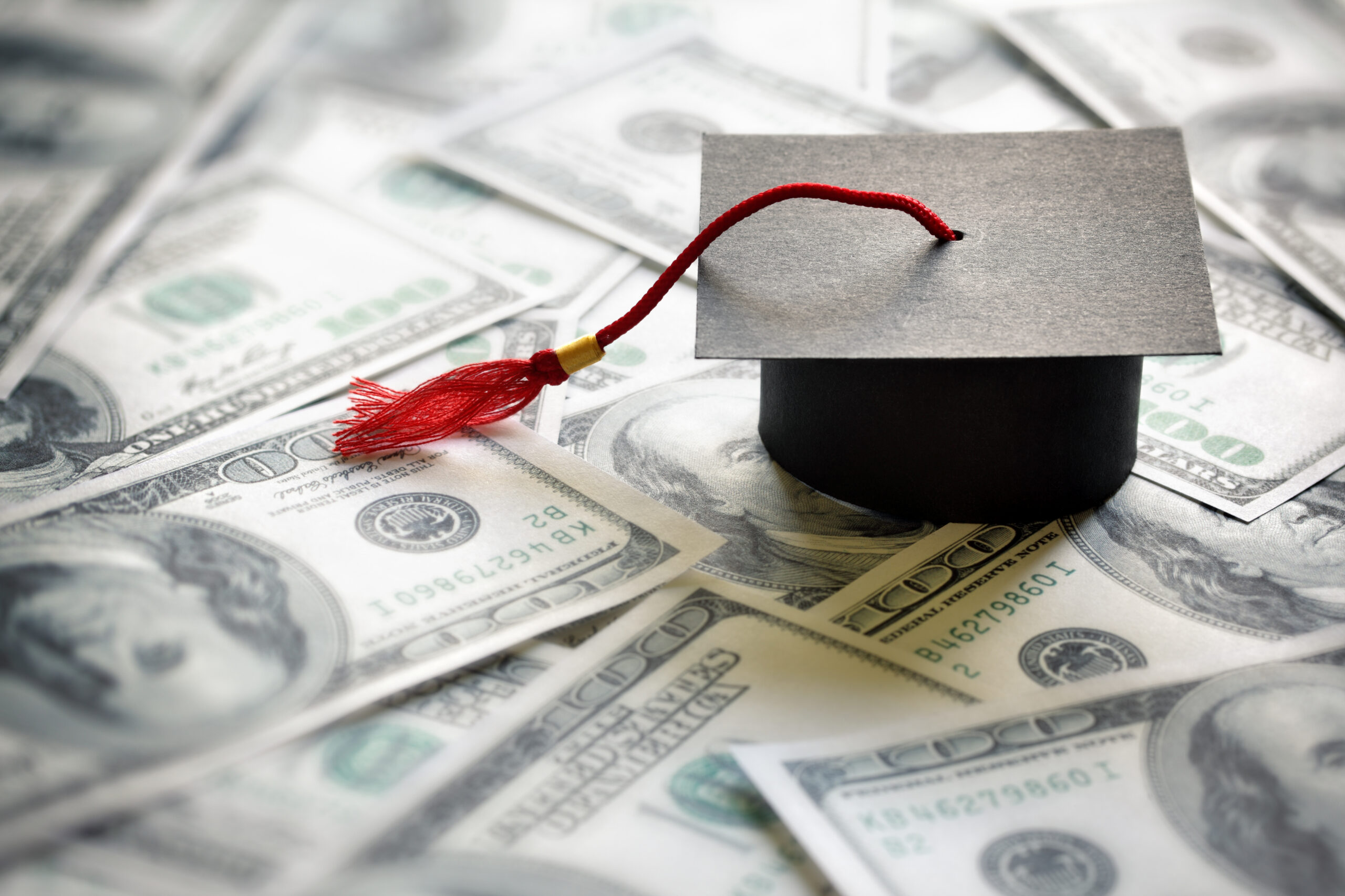A black graduation cap with a red tassel rests on a bed of scattered hundred dollar bills, symbolizing the cost of education.