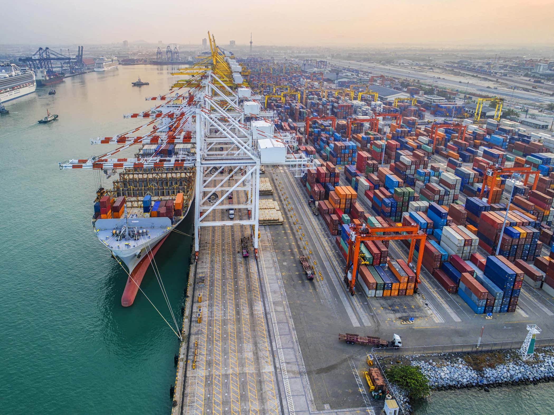 Aerial view of a busy shipping port with a large cargo ship docked, rows of colorful shipping containers stacked high, and cranes loading and unloading goods along the waterfront.