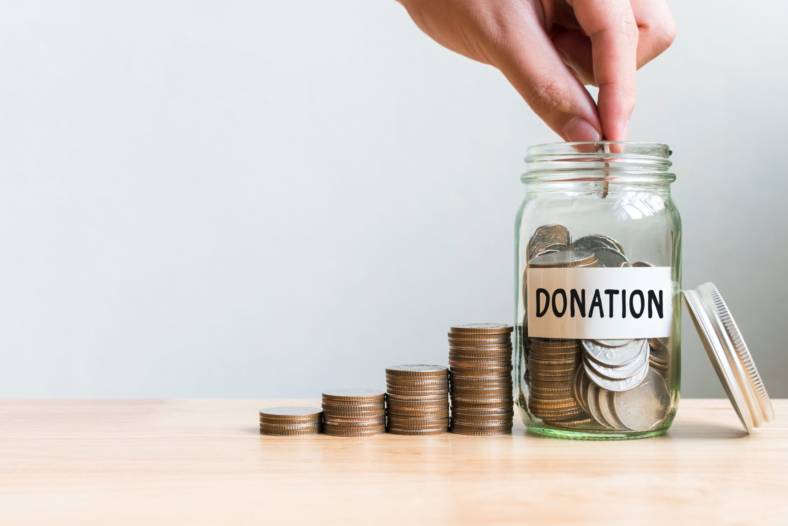 A hand drops coins into a glass jar labeled DONATION, with several stacks of coins increasing in height arranged next to the jar on a wooden surface.