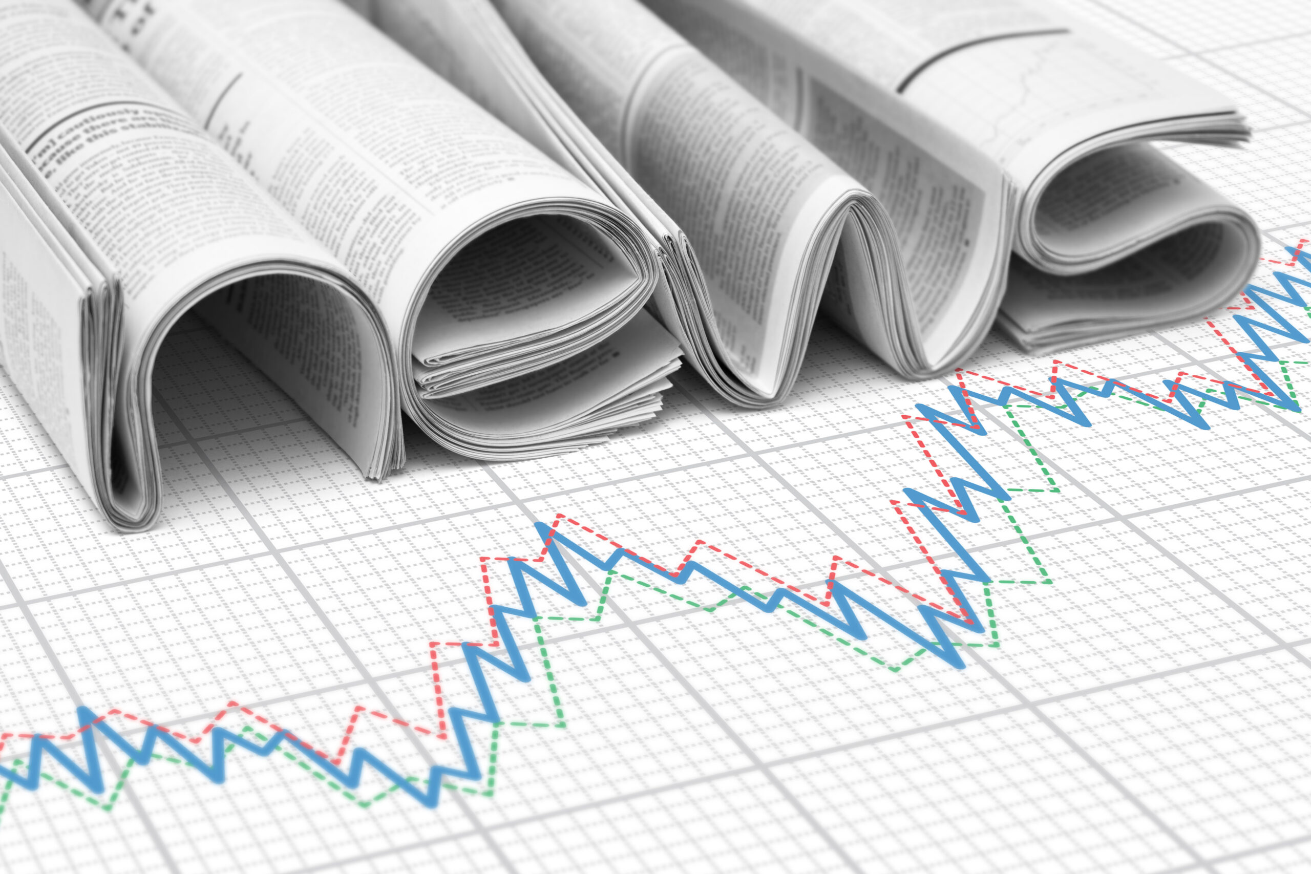 Four folded newspapers rest on a paper with multiple colored line graphs, symbolizing financial news and stock market analysis.