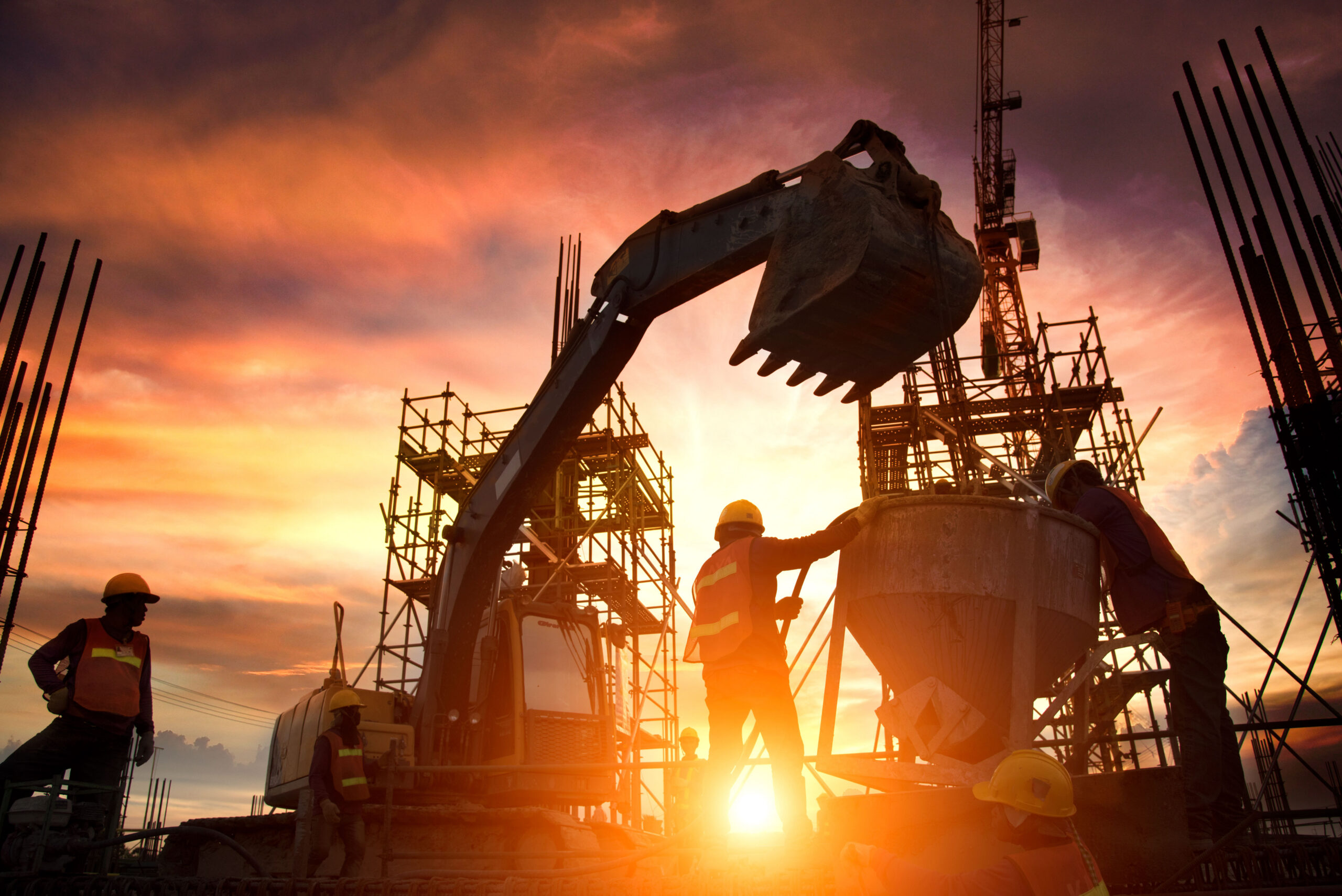 Silhouetted construction workers and an excavator work on a building site with scaffolding at sunset, creating a dramatic sky with orange and purple hues in the background.