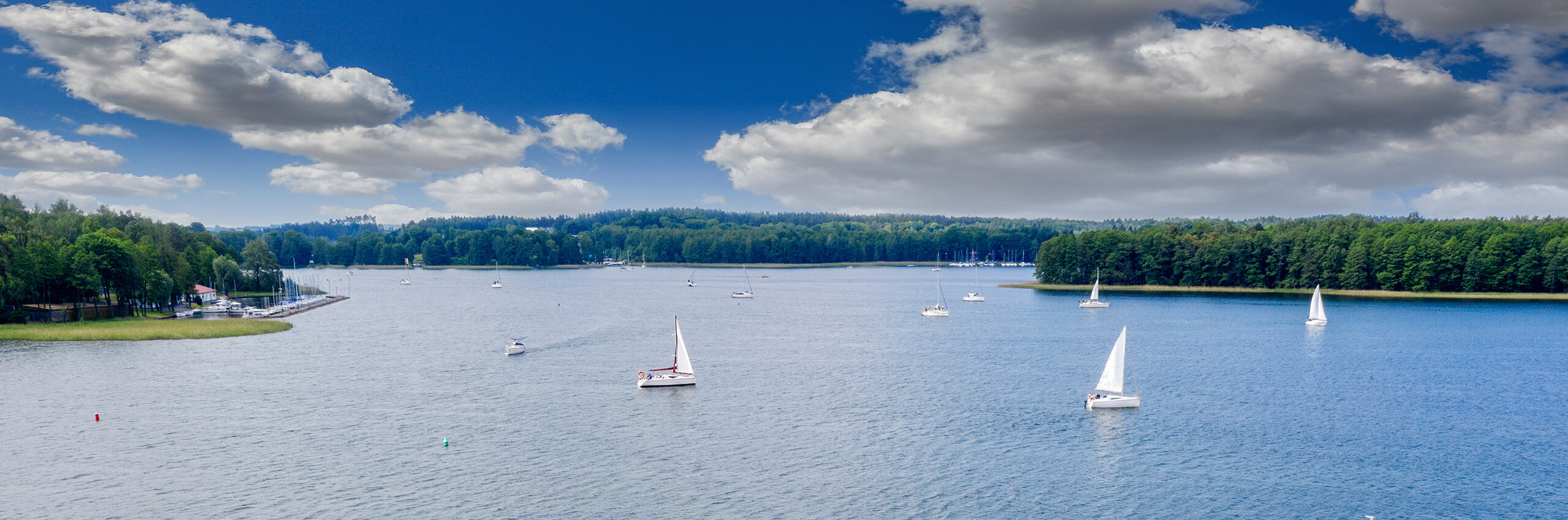 A scenic lake with several white sailboats on calm blue water, surrounded by lush green forests under a partly cloudy sky.