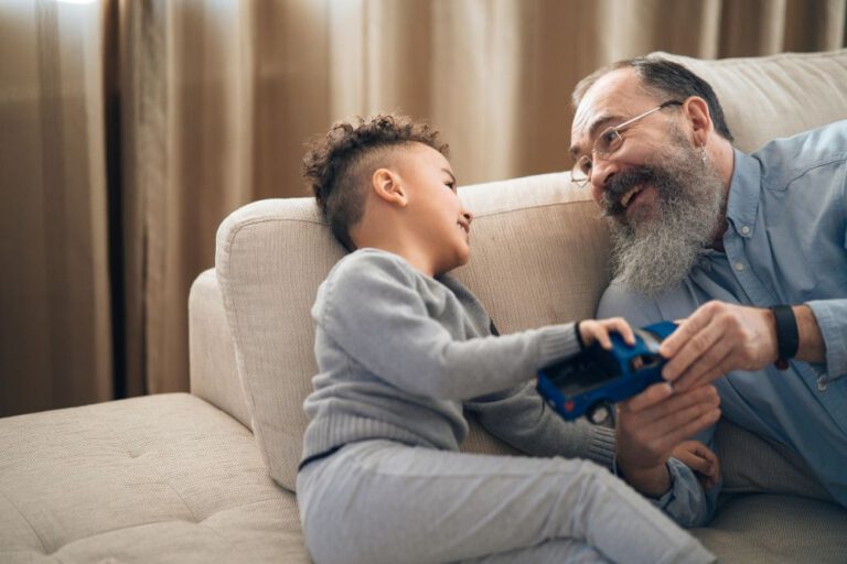 An elderly man with a beard and glasses laughs while playing with a young boy on a couch. The boy, smiling, holds a blue toy car as they enjoy their time together in a cozy living room.