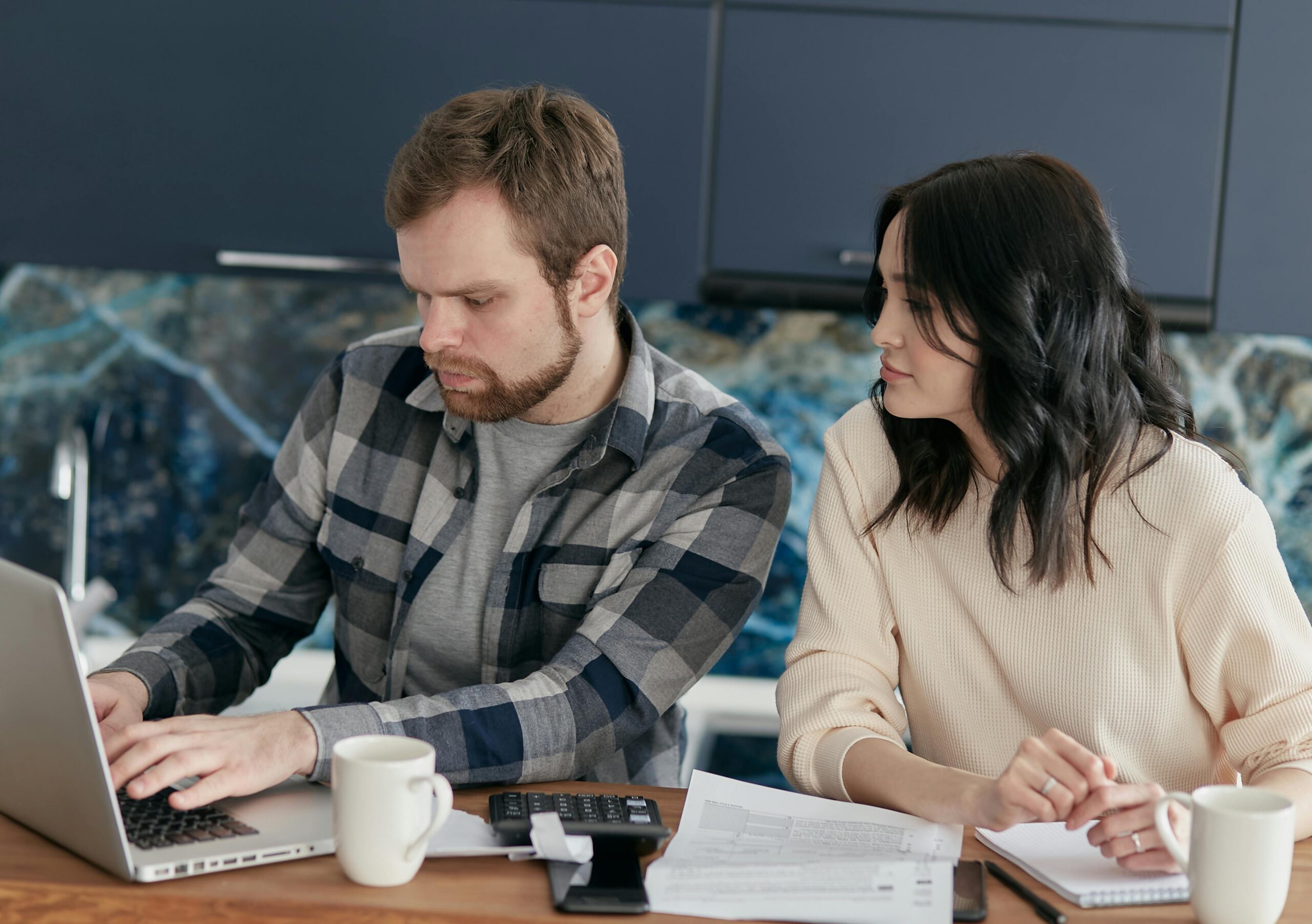 A man uses a laptop while a woman sits beside him, looking at the screen. They are seated at a table with papers, a calculator, and two mugs, suggesting they are working on finances in a kitchen setting.