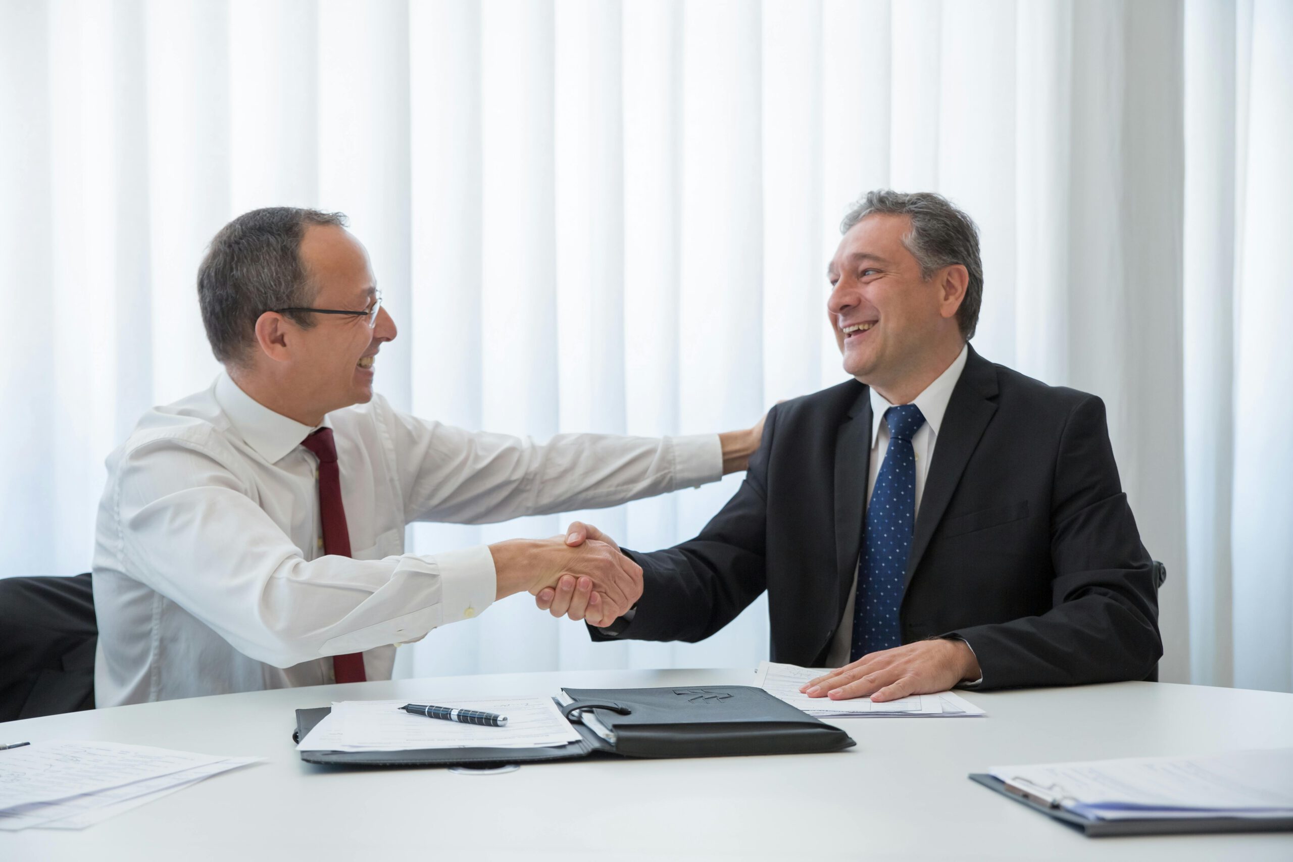 Two men in business attire sit at a table, smiling and shaking hands, with documents and a pen in front of them, suggesting a successful business agreement or partnership.