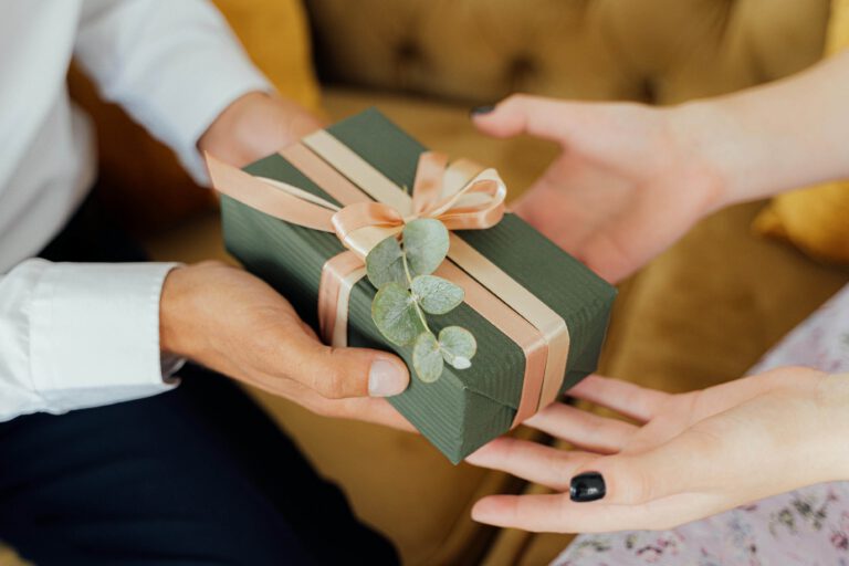 A person hands a green gift box with a peach ribbon and sprig of eucalyptus to another person, who is reaching out to receive it. They are seated on a tan couch.