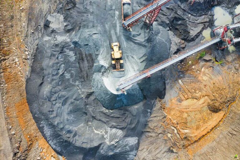 Aerial view of a mining site with a large yellow excavator moving gray rocks, surrounded by conveyors and earth, creating circular patterns in the landscape.