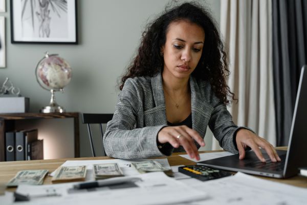 A woman in a gray blazer sits at a desk using a laptop and calculator, with cash, papers, and bills spread out in front of her, suggesting she is managing finances or budgeting.