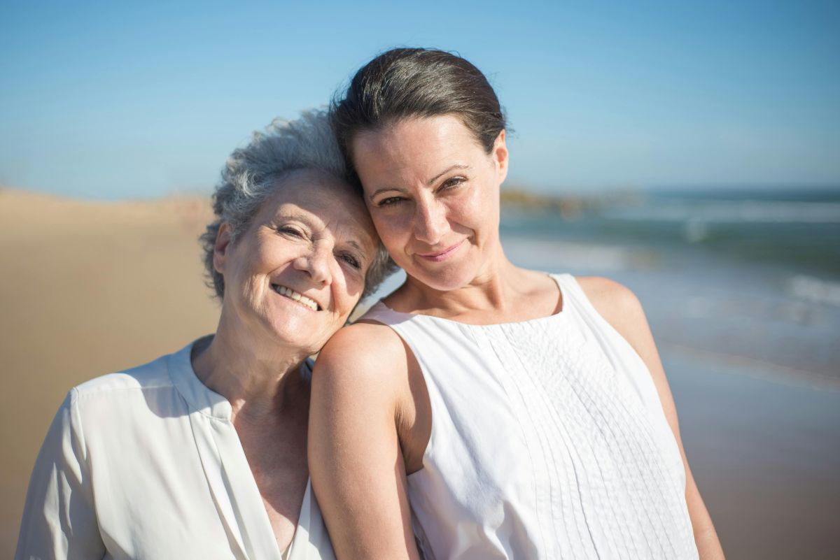 Two women stand close together on a sunny beach; one older with gray hair smiles while leaning her head on the younger woman, who has brown hair and is also smiling. Both wear white tops. The sea and blue sky are in the background.