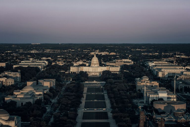Aerial view of the U.S. Capitol building at sunset, surrounded by trees and government buildings, with the National Mall stretching out in the foreground. The sky is clear with a purple and blue gradient.