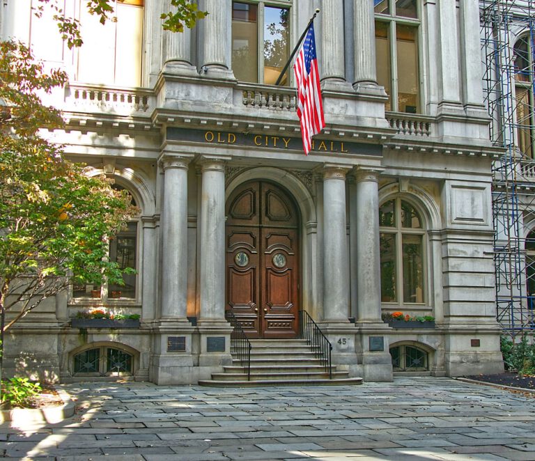Grand stone entrance of Old City Hall with large wooden doors, tall columns, an American flag above the steps, and sunlight illuminating the historic building’s facade. Trees and potted plants frame the entrance.