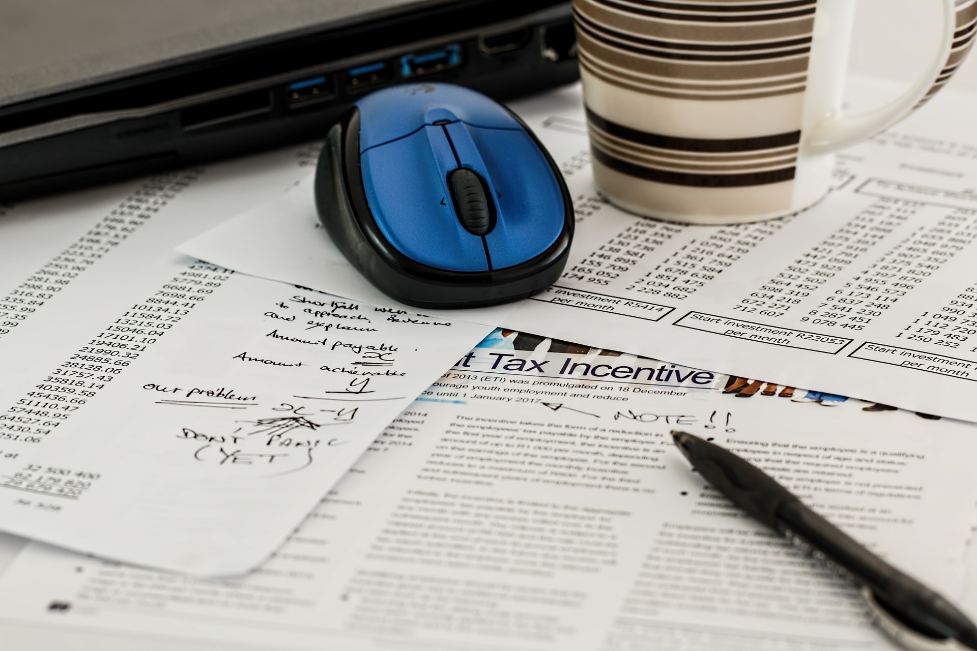 A blue computer mouse, a striped coffee cup, and a pen rest on scattered documents and notes, including a tax incentive bill and handwritten calculations, on a desk with a partially visible laptop.