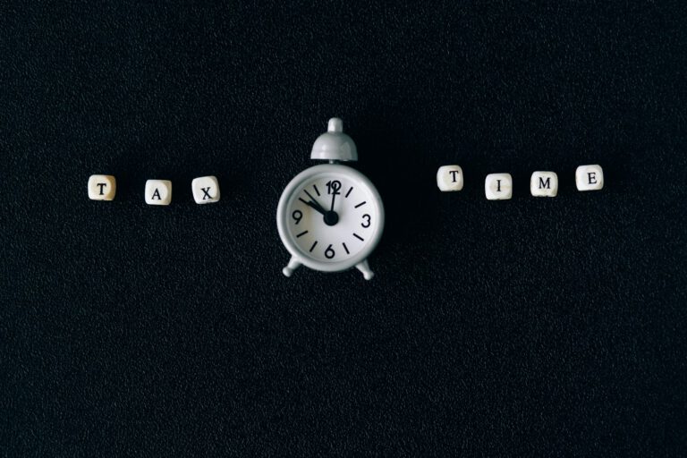 A small white clock centered on a dark surface with lettered beads spelling TAX on the left and TIME on the right.