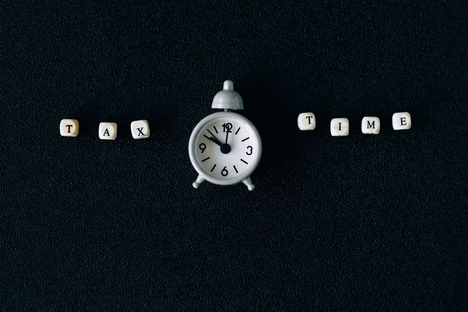 A small white clock centered on a dark surface with lettered beads spelling TAX on the left and TIME on the right.