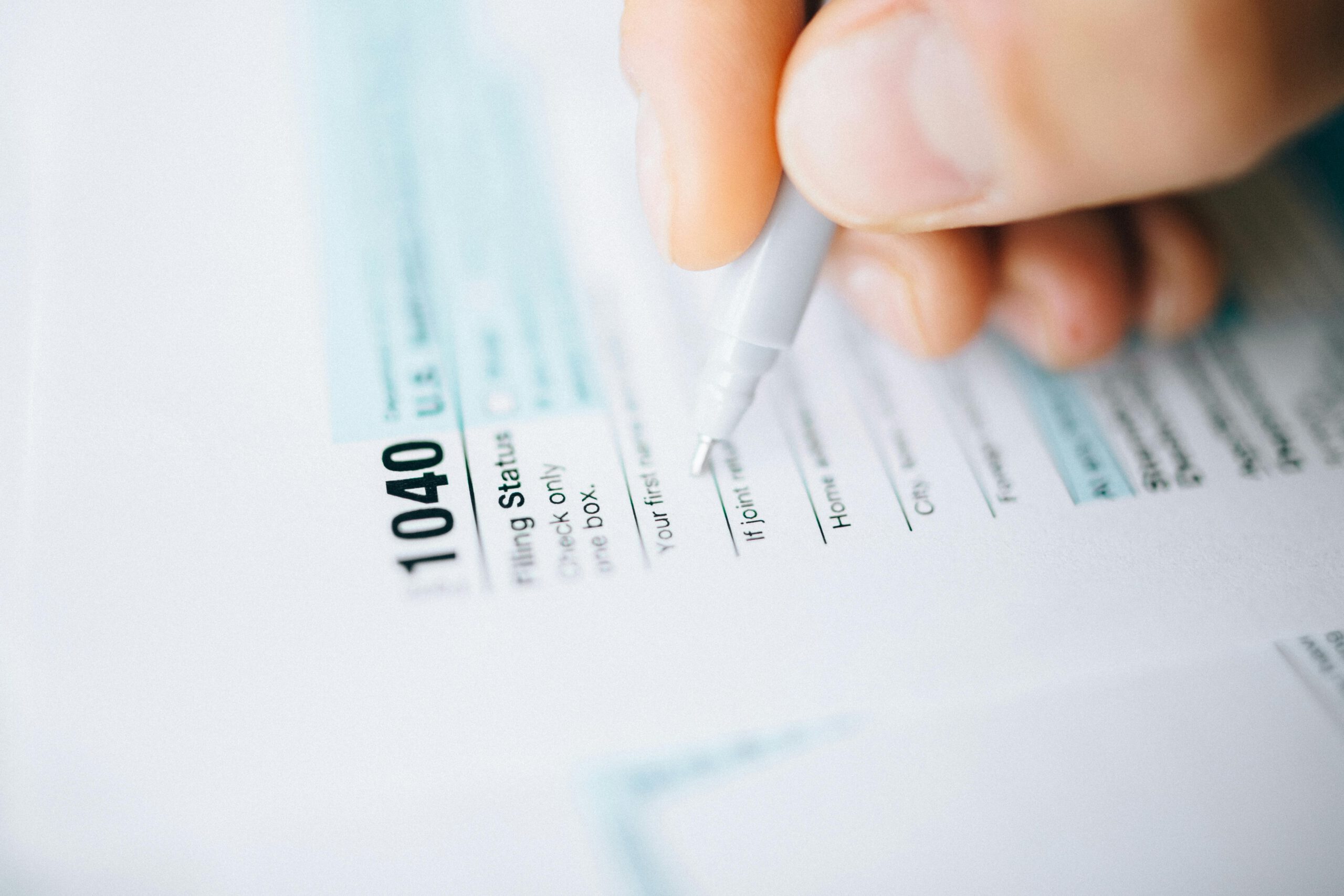 A close-up of a hand holding a pen, filling out a U.S. IRS Form 1040 tax document. The form details and personal information fields are partially visible.