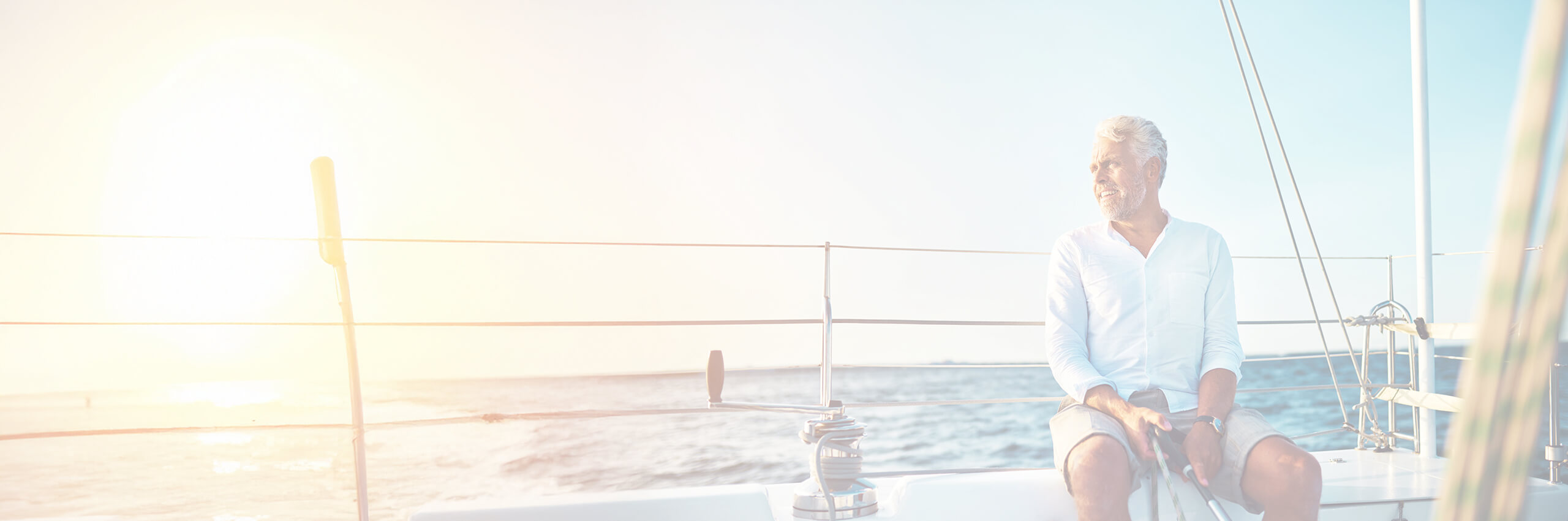 An older man with gray hair sits relaxed on a sailboat in sunlight, looking out at the ocean. The water and sky are bright and calm, creating a peaceful, summery atmosphere.