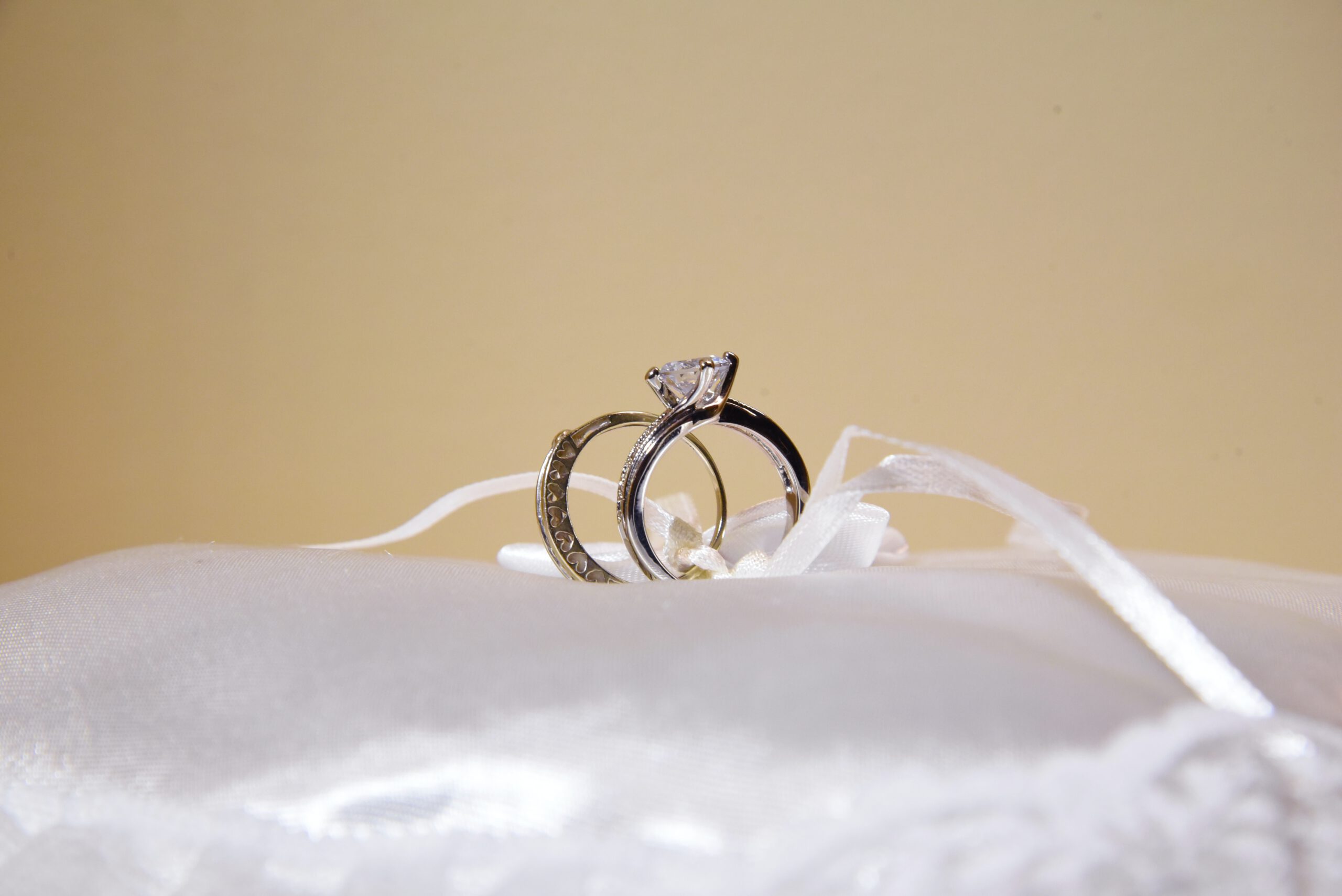 Two silver rings, one with a diamond, are tied together with white ribbon and placed on a white satin cushion, against a light beige background.
