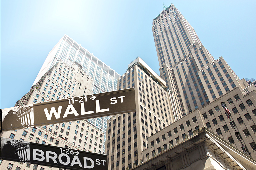 Street signs for Wall Street and Broad Street with tall skyscrapers in the background under a clear blue sky, representing the Financial District in New York City.