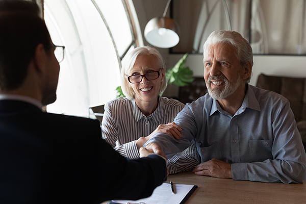 An older couple sits at a table, smiling and shaking hands with a person in a suit across from them, suggesting a successful meeting or agreement in a bright, modern office setting.