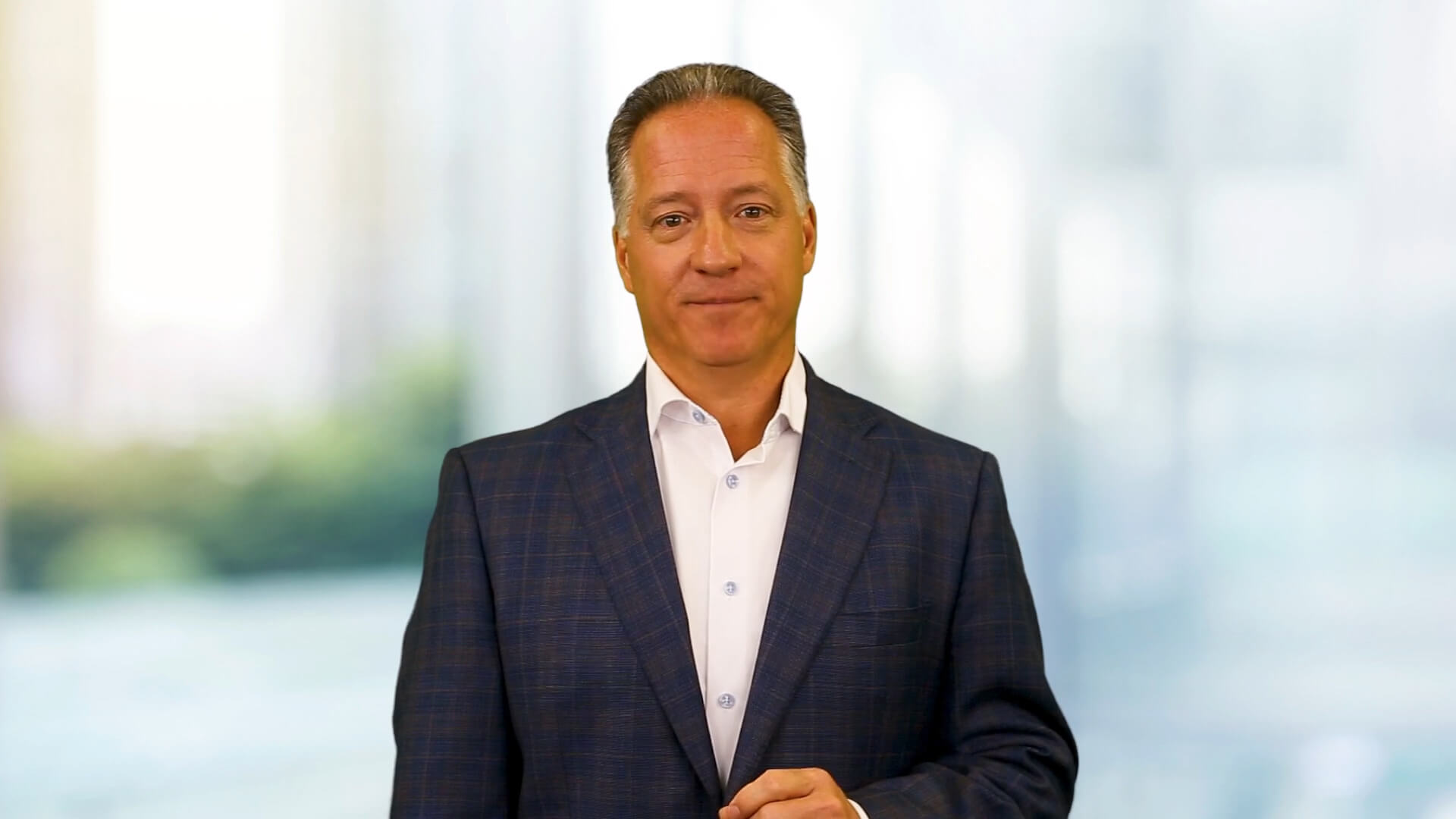 A middle-aged man in a dark plaid suit and white shirt stands indoors in front of a blurred background, looking at the camera with a neutral expression.