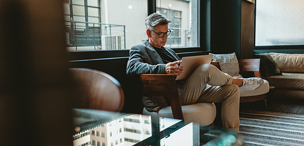 A man with gray hair and glasses sits in a modern lounge, focused on a tablet. He wears a blazer, light pants, and sneakers, reviewing his concentrated stock position as natural light streams through large windows behind him.